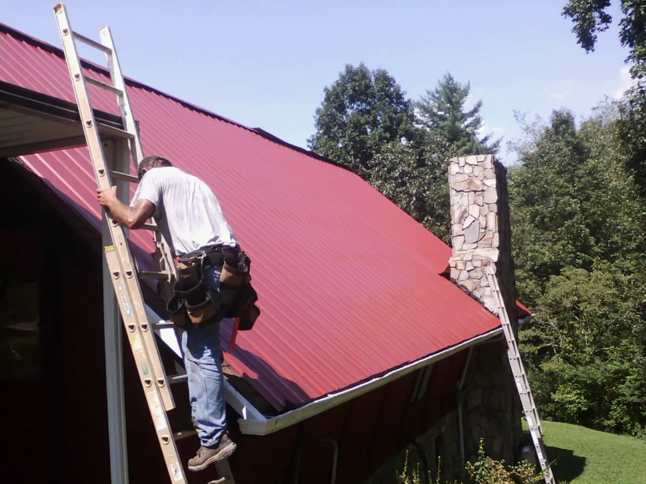 Roofer on a ladder beside a red metal roof and stone chimney — Soffit & Fascia Repair by Roofing Brothers in Amherst